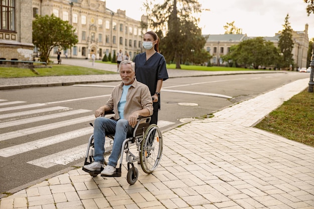 Young nurse in protective face mask assisting senior handicapped man in wheelchair during a walk in