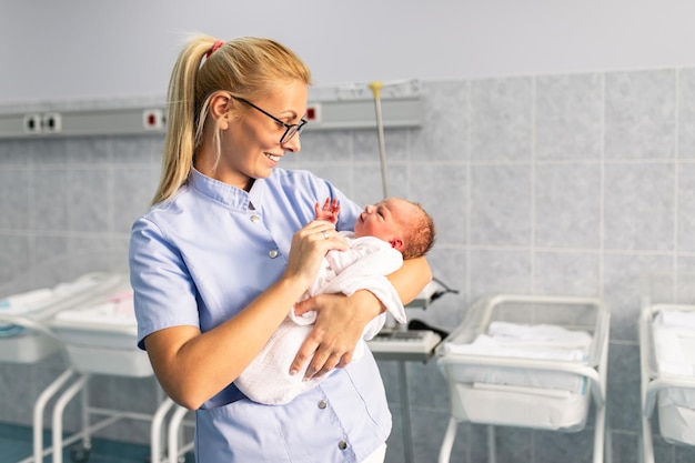 Young nurse standing in maternity ward and holding newborn baby in her arms. After birth concept.