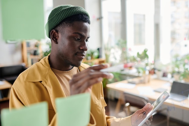 Young office worker in casualwear looking at tablet screen by noticeboard