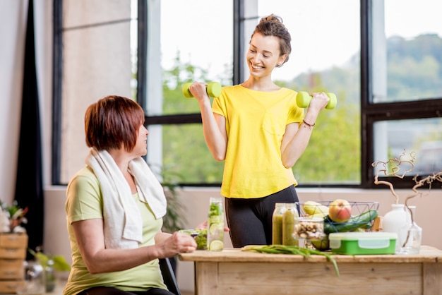 Photo young and older women exercising with dumbbells and eating healthy food indoors on the window background