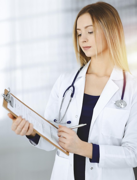 Young optimistic woman-doctor is holding a clipboard in her hands, while standing in a sunny clinic. Portrait of friendly female physician with a stethoscope. Perfect medical service in a hospital. Me