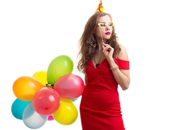 Young optimistic woman with celebrating hat holding many colored balloons and card stick of glasses