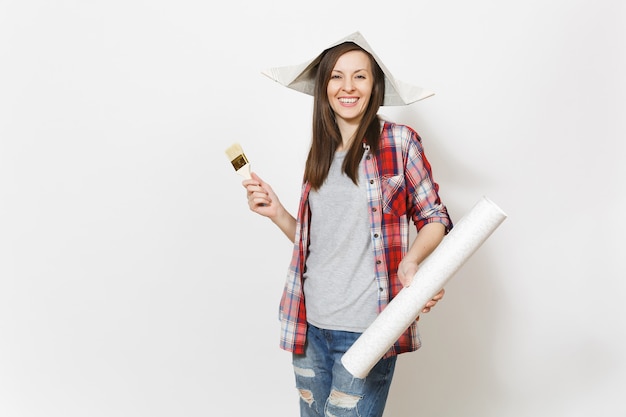 Photo young overjoyed beautiful woman in casual clothes and newspaper hat holding paint brush and wallpaper roll isolated on white background. instruments for renovation apartment room. repair home concept.