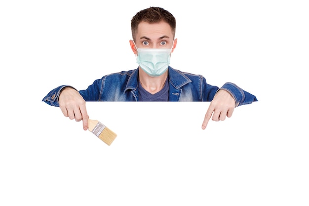 A young painter in a denim jacket with a medical mask on his face holds a paint brush in his hand