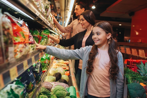 Young parents and daughter in grocery store. They stand in front of salad shelf and pick it up. People smile. Happy family together.