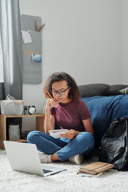 Young pensive cross-legged female student sitting on the floor by bed in front of laptop and looking through her notes in copybook in bedroom