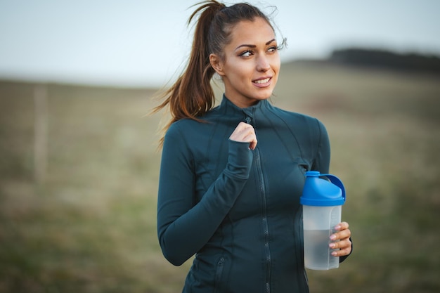 Young pensive female runner is drinking water and taking a break after jogging in mountains. Looking away.