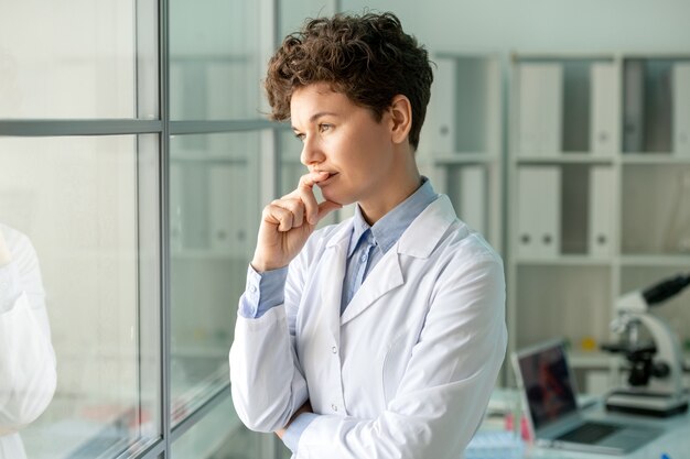 Young pensive female scientist or laboratory worker in whitecoat standing by glass wall against her workplace with laptop and microscope