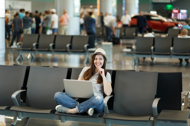 Young pensive traveler tourist woman in hat sitting with crossed legs working on laptop wait in lobby hall at international airport