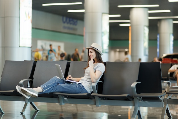 Young pensive traveler tourist woman in hat working on laptop while waiting in lobby hall at international airport