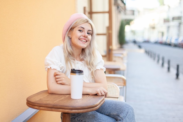 Young pensive woman in beret smiling looking at the camera while sitting at a table in outdoor cafe on European street