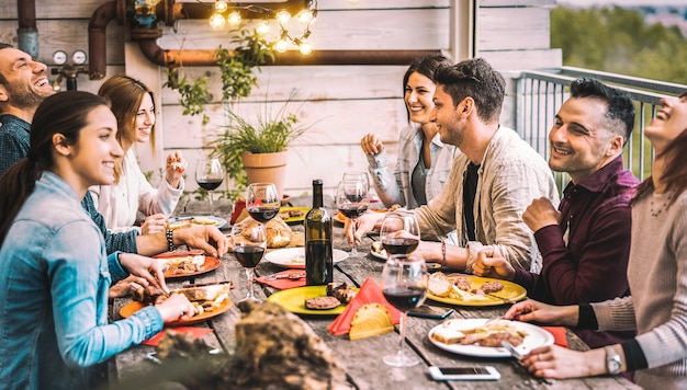 Young people dining and having fun drinking red wine together on balcony rooftop dinner party - Happy friends eating bbq food at restaurant patio - Millannial life style concept