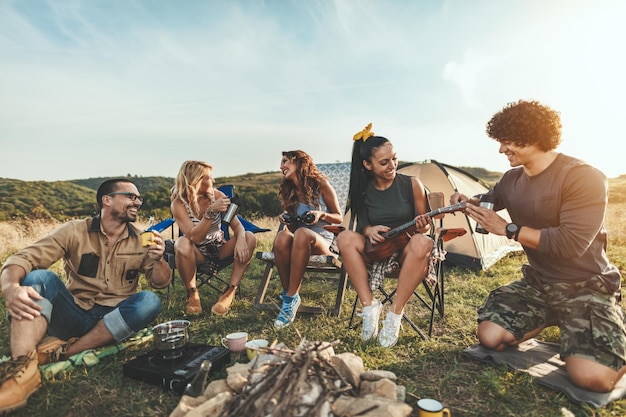 Young people have a good time in camp in nature. They're resting, laughing and singing by ukulele music, happy to be together.