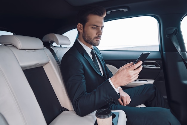 Young perfectionist. Handsome young man in full suit using his smart phone while sitting in the car