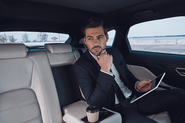 Young perfectionist. Handsome young man in full suit working using digital tablet while sitting in the car