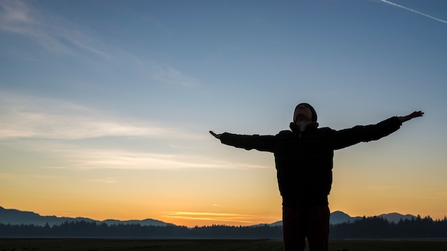 Young person celebrating the sunrise or sunset standing with outstretched arms silhouetted against a colorful orange sky.