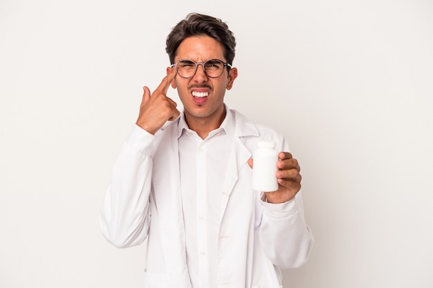 Young pharmacist mixed race man holding pills isolated on white background showing a disappointment gesture with forefinger.