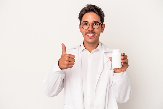 Young pharmacist mixed race man holding pills isolated on white background smiling and raising thumb up