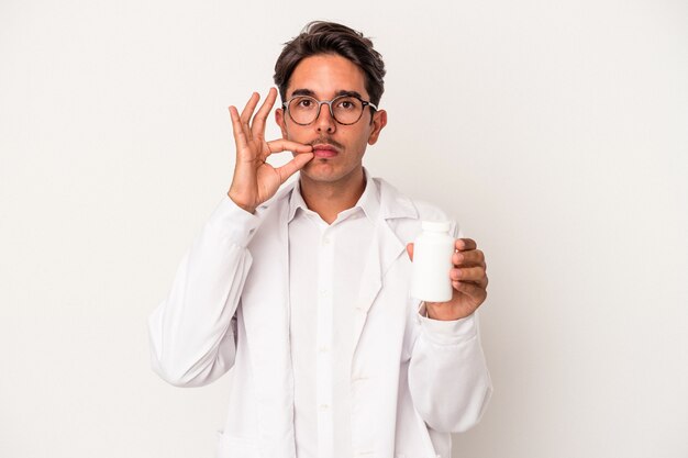 Young pharmacist mixed race man holding pills isolated on white background with fingers on lips keeping a secret.