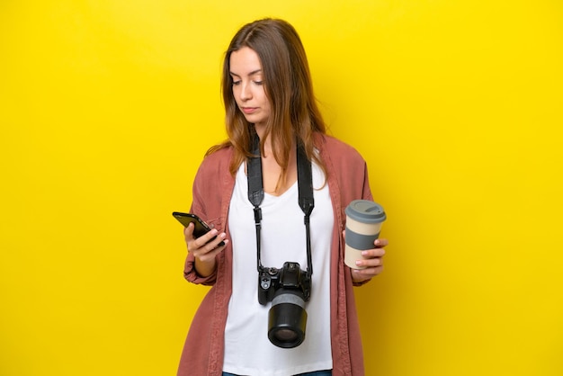 Young photographer caucasian woman isolated on yellow background holding coffee to take away and a mobile