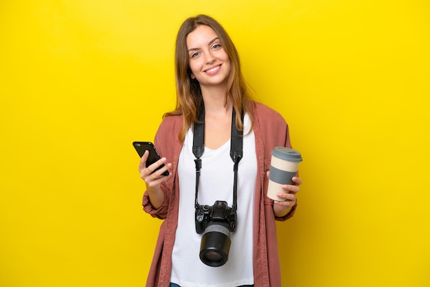 Young photographer caucasian woman isolated on yellow background holding coffee to take away and a mobile