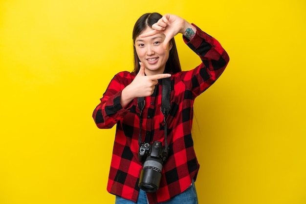 Young photographer Chinese woman isolated on yellow background focusing face Framing symbol