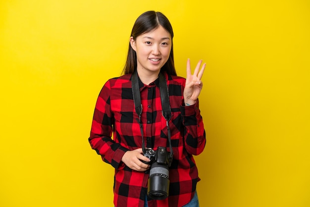 Young photographer Chinese woman isolated on yellow background happy and counting three with fingers