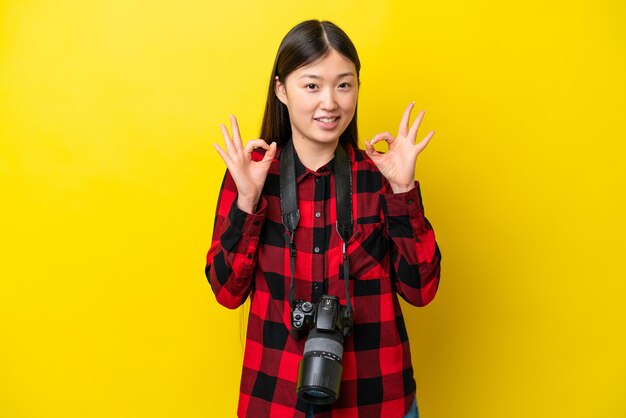 Young photographer Chinese woman isolated on yellow background showing an ok sign with fingers