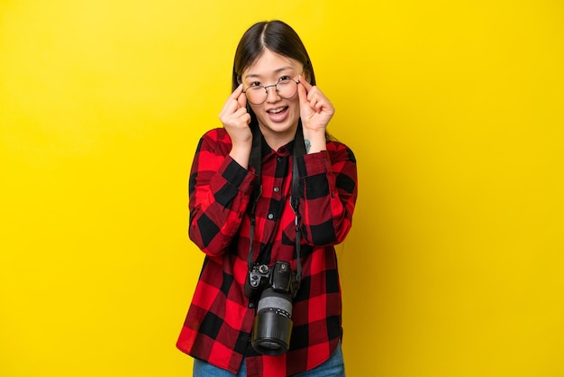 Young photographer Chinese woman isolated on yellow background with glasses and surprised