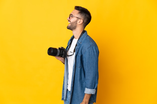 Young photographer girl isolated on yellow background laughing in lateral position