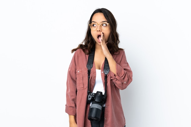 Young photographer woman over isolated white wall whispering something with surprise gesture while looking to the side