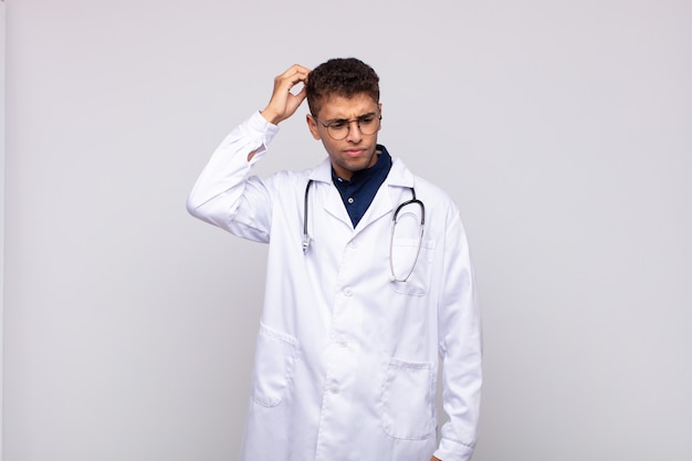 Young physician man feeling puzzled and confused, scratching head and looking to the side