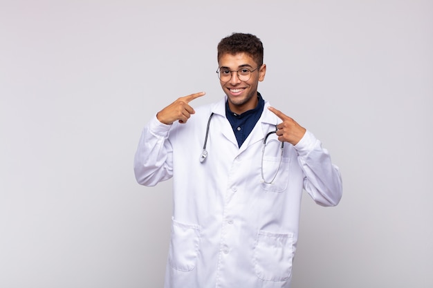 Young physician man smiling confidently pointing to own broad smile, positive, relaxed, satisfied attitude