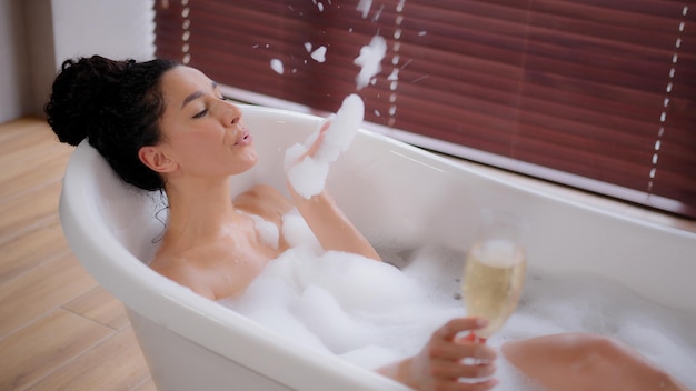 Young playful woman relaxing in bath with bubbles holding glass champagne enjoying blowing on foam