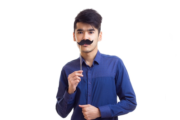 Young pleasant man with dark hair in blue shirt holding cardboard stick of moustache in studio