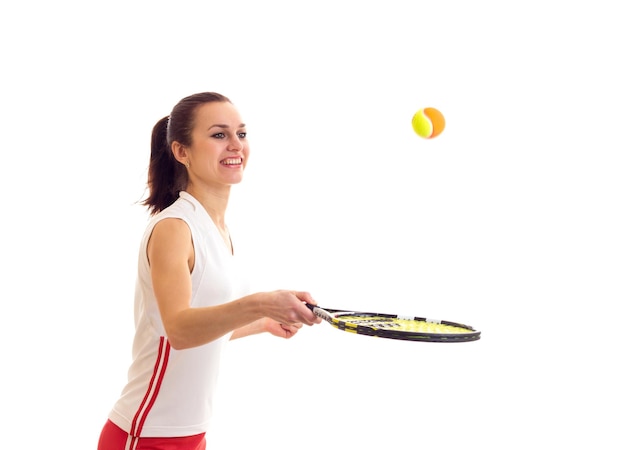 Young pleasant woman in white sports shirt and red skirt with chestnut ponytail playing with tennis raquet and yellow ball on white background in studio