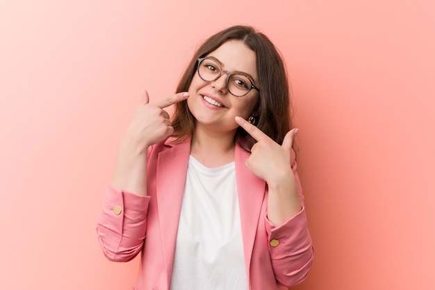 Young plus size business caucasian woman smiles, pointing fingers at mouth.