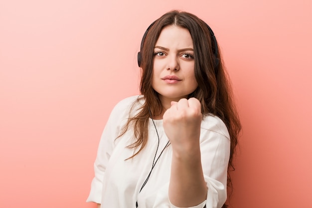 Young plus size curvy woman listening music with headphones showing fist, aggressive facial expression.