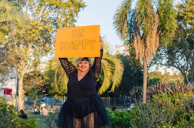 Young plus size woman in the park with a yellow sign that says be happy