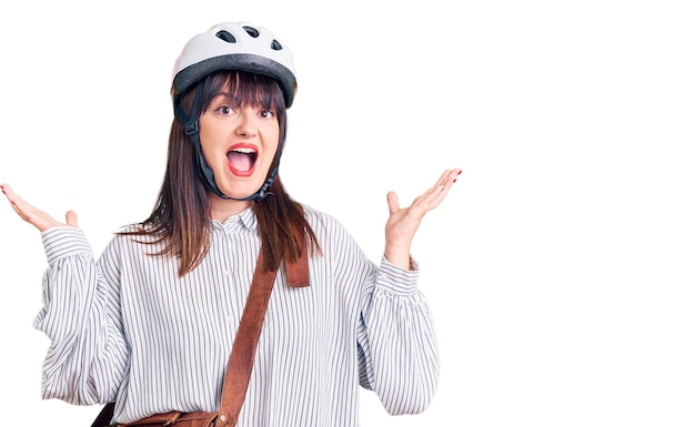 Young plus size woman wearing bike helmet and leather bag celebrating victory with happy smile and winner expression with raised hands