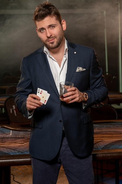 Young poker player with glass of drink and pair of aces standing in casino