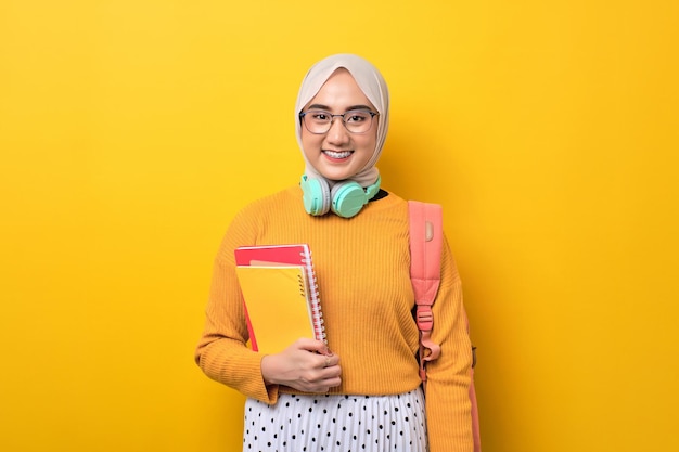 Young positive Asian student girl with backpack holding notebook smiling at camera isolated over yellow background