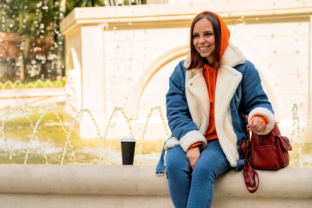 Young positive beautiful woman in casual warm clothes with coffee in paper cup sitting on fountain in city park