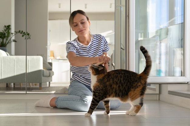 Young positive Caucasian woman is doing grooming procedures for kitten sits on floor in apartment