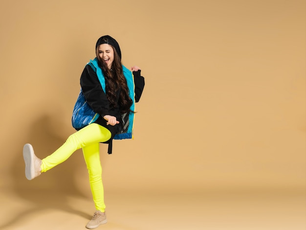 Young positive girl in yellow leggings and a warm vest makes a step with her foot on a pastel orange.