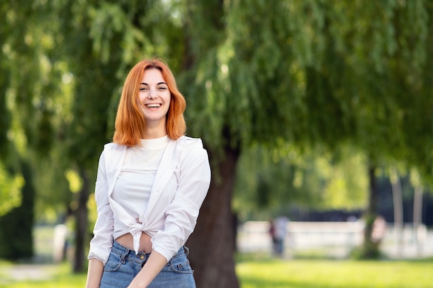 Young positive redhead girl in casual clothes standing in summer park.