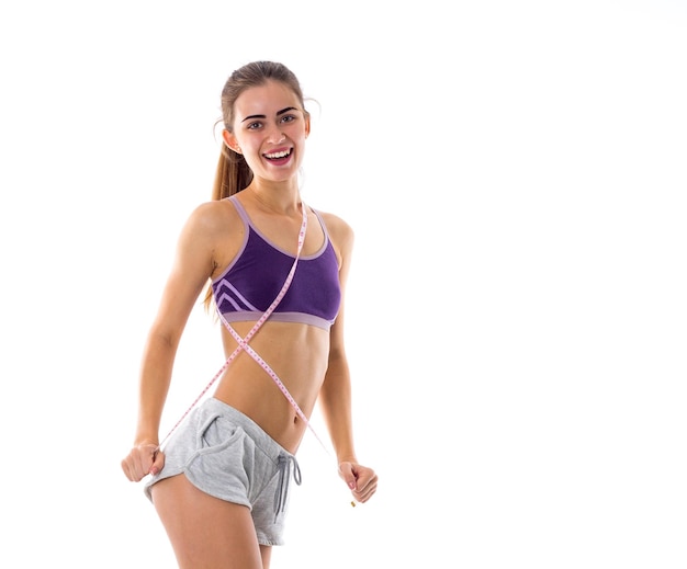 Young positive woman in purple sports top with ponytail using centimeter on white background in studio