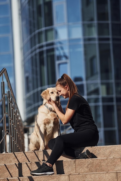 Young positive woman sitting on stairs with her dog when have a walk outdoors near business building