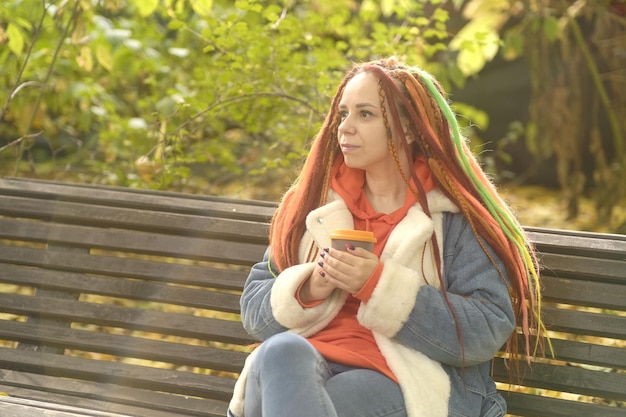 Young positive woman with ginger dreadlocks drinking coffee looking away sitting on park bench in golden autumn