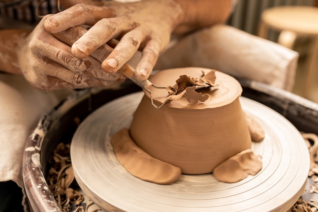 Young potter using special handtool to process bottom of new clay item while bending over pottery wheel